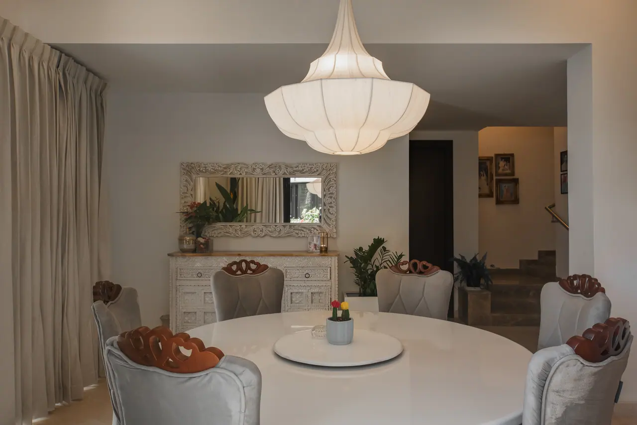 Dining table under a large fabric pendant, with a carved white sideboard and mirror beyond and steps leading up at right