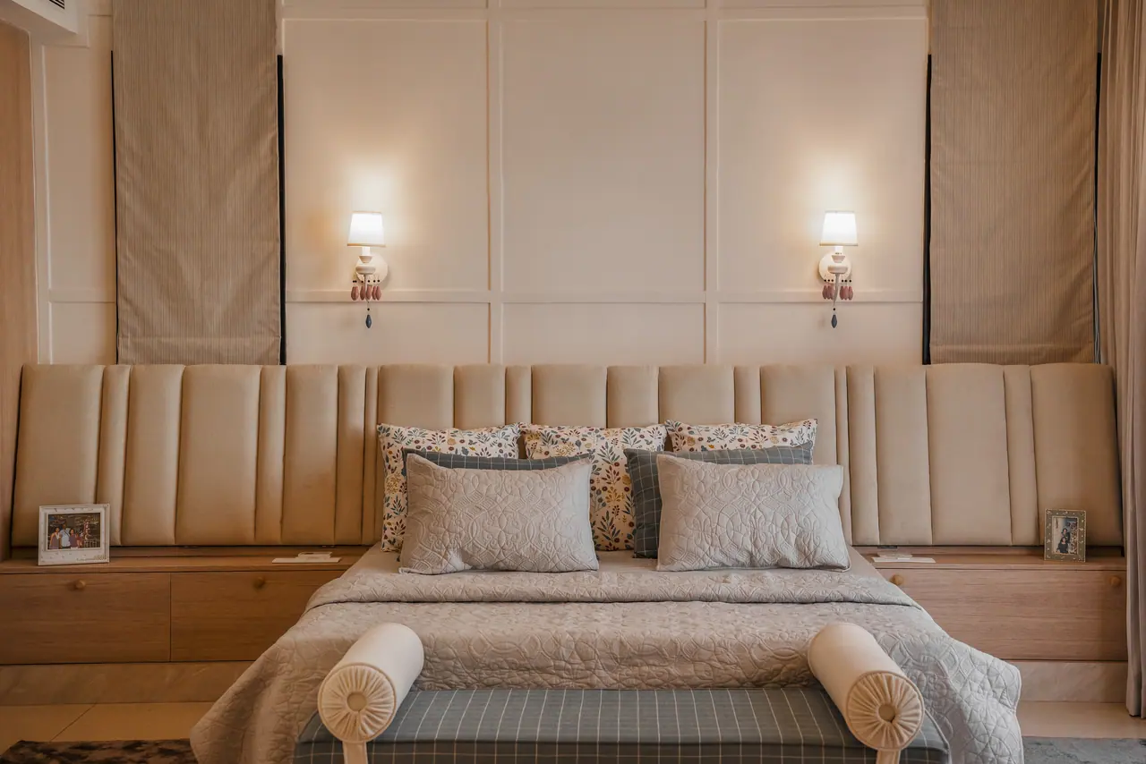 Channel-tufted headboard spanning wood bedside units, with panelled wall, twin sconces, Roman blinds and a checked bench