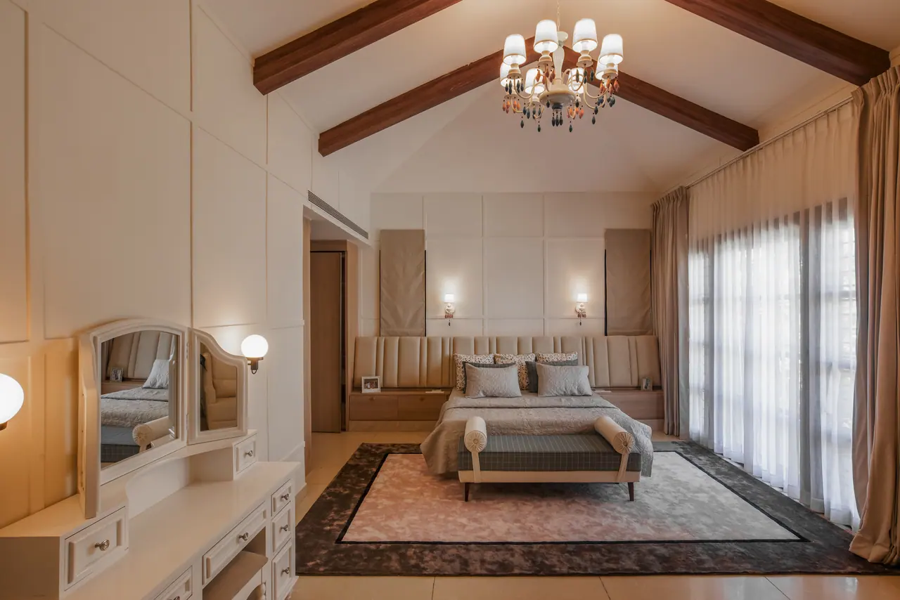 Bedroom with a vaulted beamed ceiling, chandelier, channel-tufted headboard wall, rose rug, vanity mirror and sheer curtains