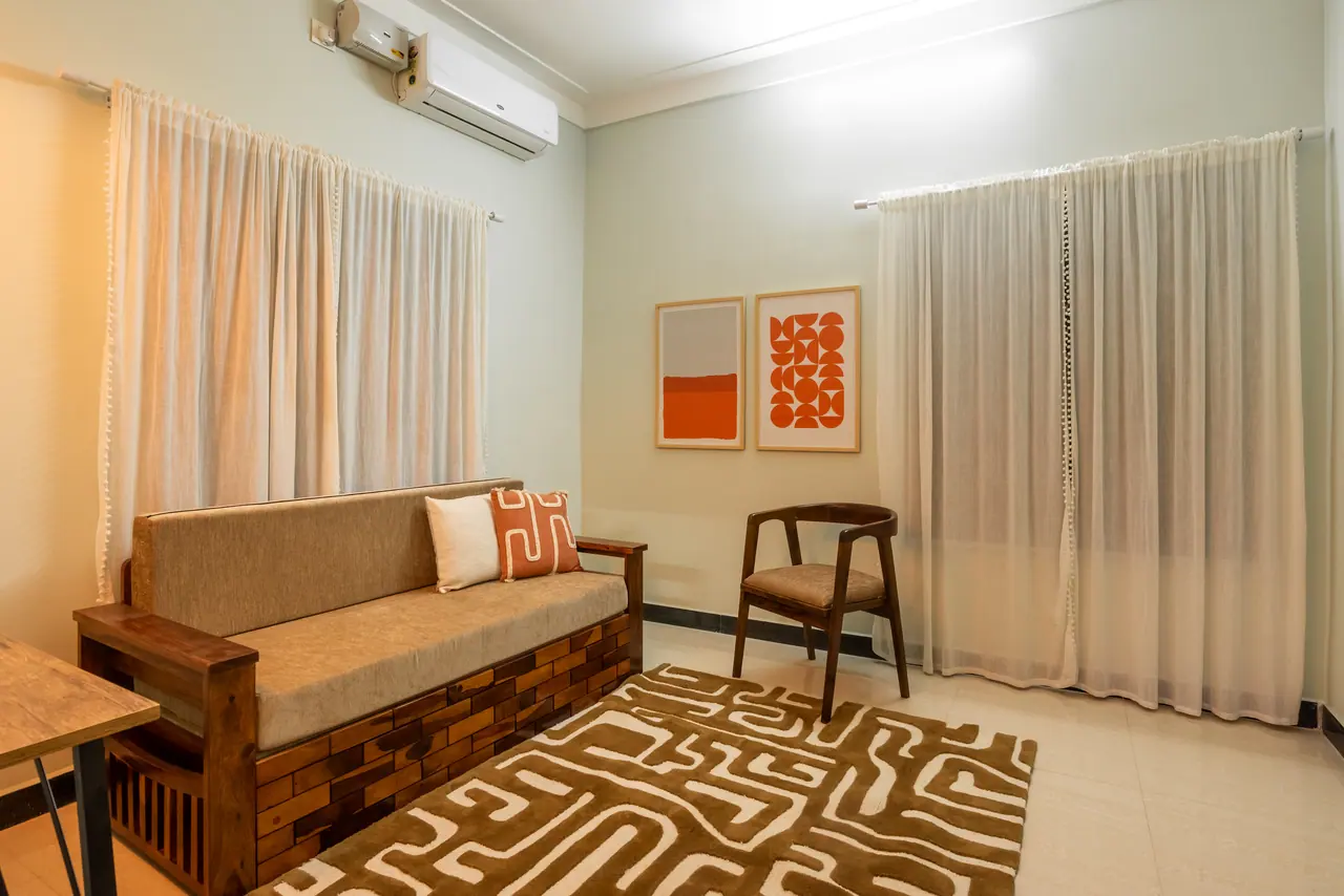 Sitting room with a wooden sofa on a block-pattern base, sheer curtains, framed orange prints, wooden armchair and maze rug