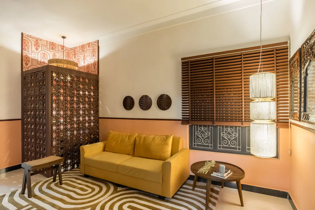 Living area with a yellow sofa, wood blinds over a scrolled window grille, tiered fabric pendant and a round wooden table