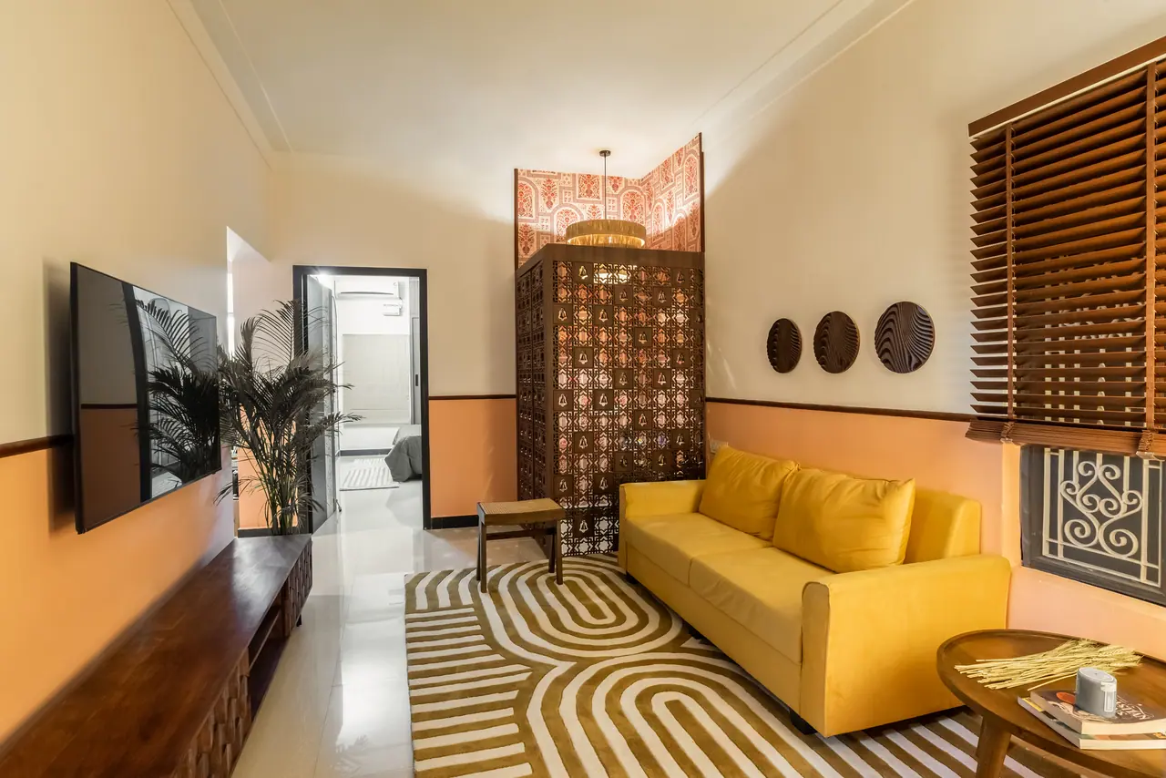 Long living room with a wall-mounted TV over a low wood console, yellow sofa, looped-stripe rug and a lattice-screened niche