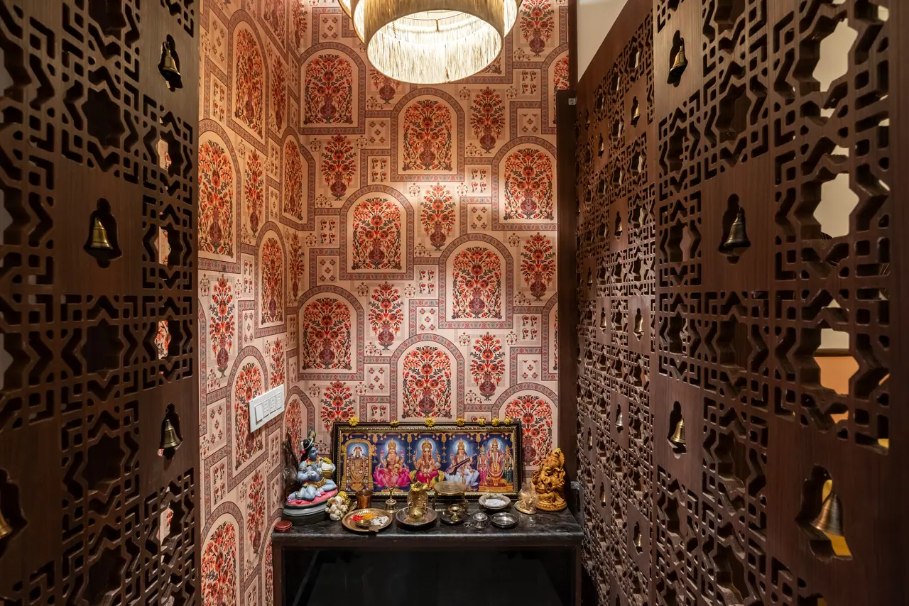 Prayer niche framed by brass-belled lattice panels, with arched floral wallpaper, a fringed pendant and idols on a counter