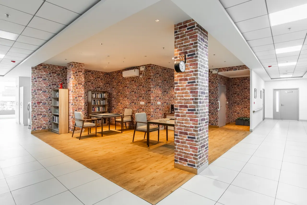 Reading corner with brick-pattern walls and column, bookshelves, tables and chairs on wood flooring in a white tiled hall