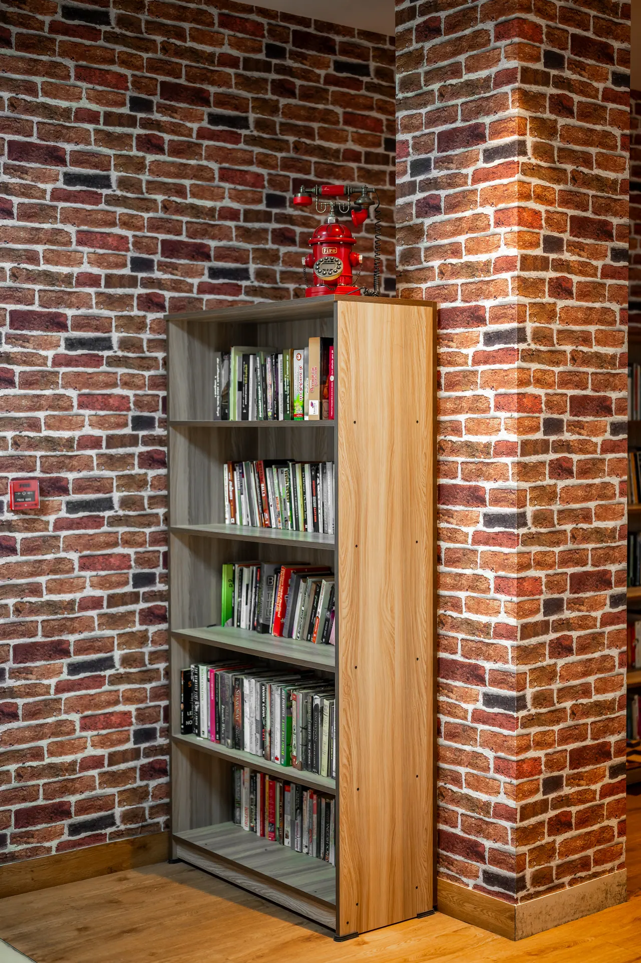 Wooden bookshelf filled with books against a red brick-pattern wall, with a red vintage-style telephone on top