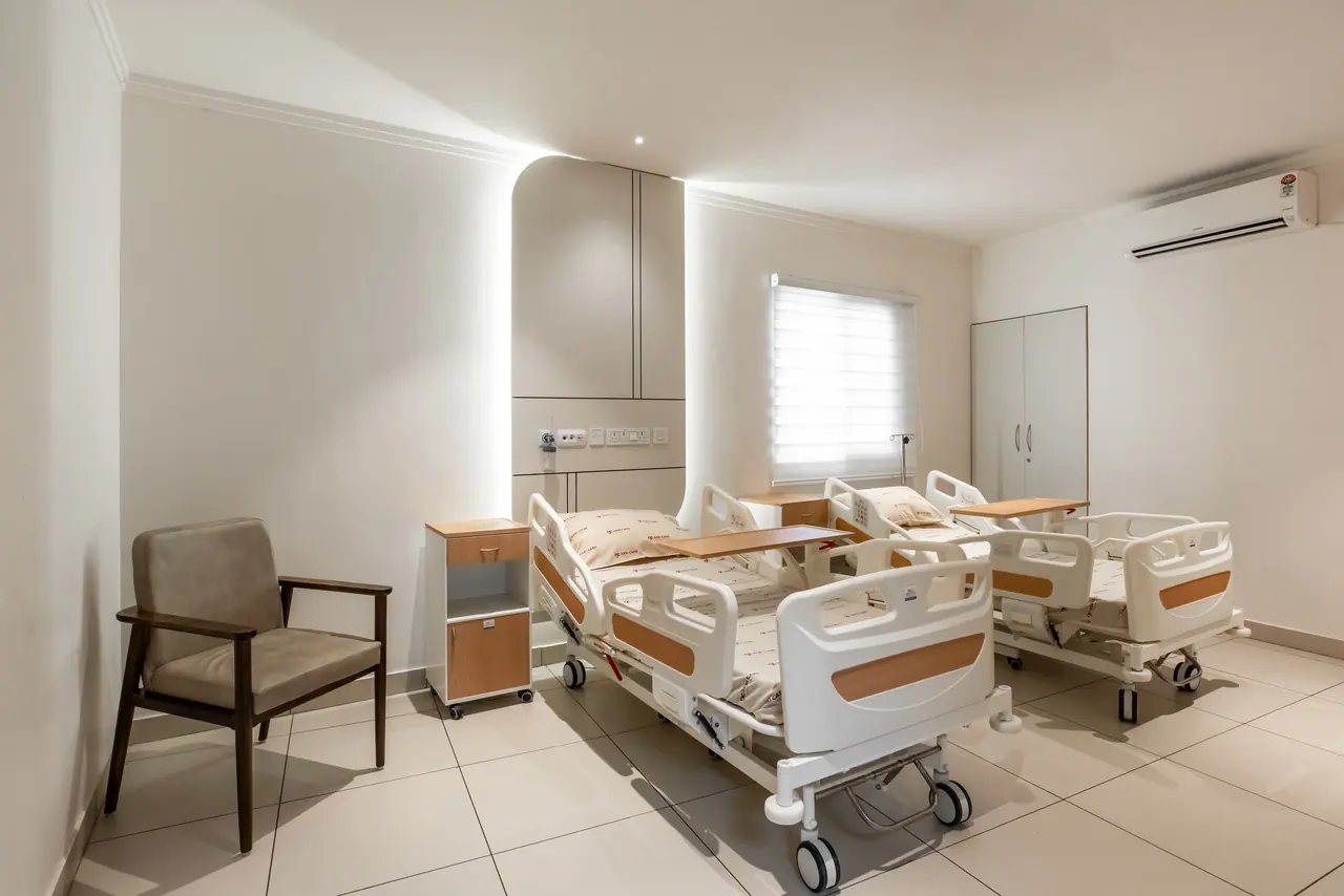 Twin patient room with adjustable hospital beds, backlit arched headwall panel, wooden armchair, bedside cabinet and blind