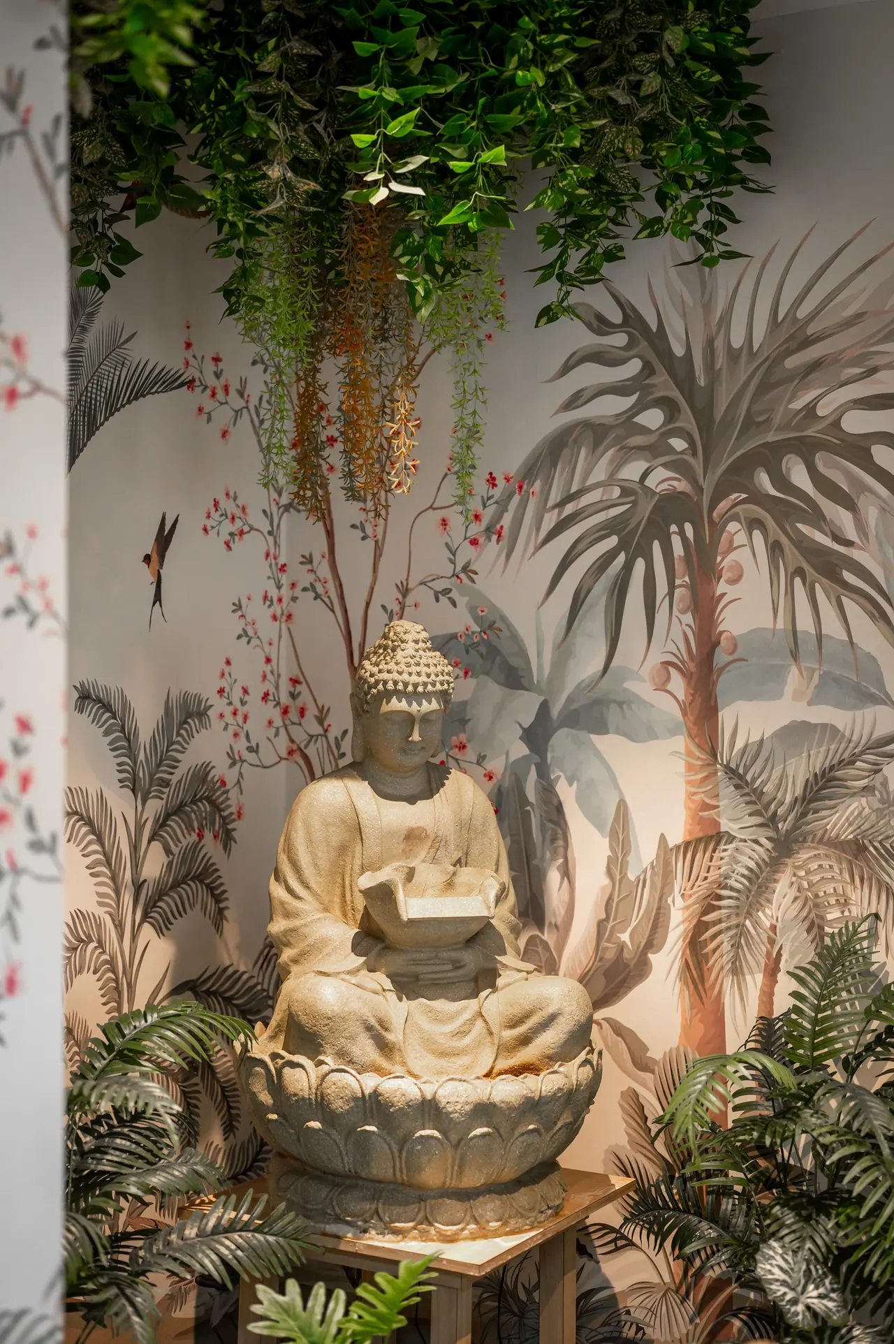 Cream seated Buddha fountain on a lotus base, framed by hanging greenery, ferns and a tropical wall mural with birds