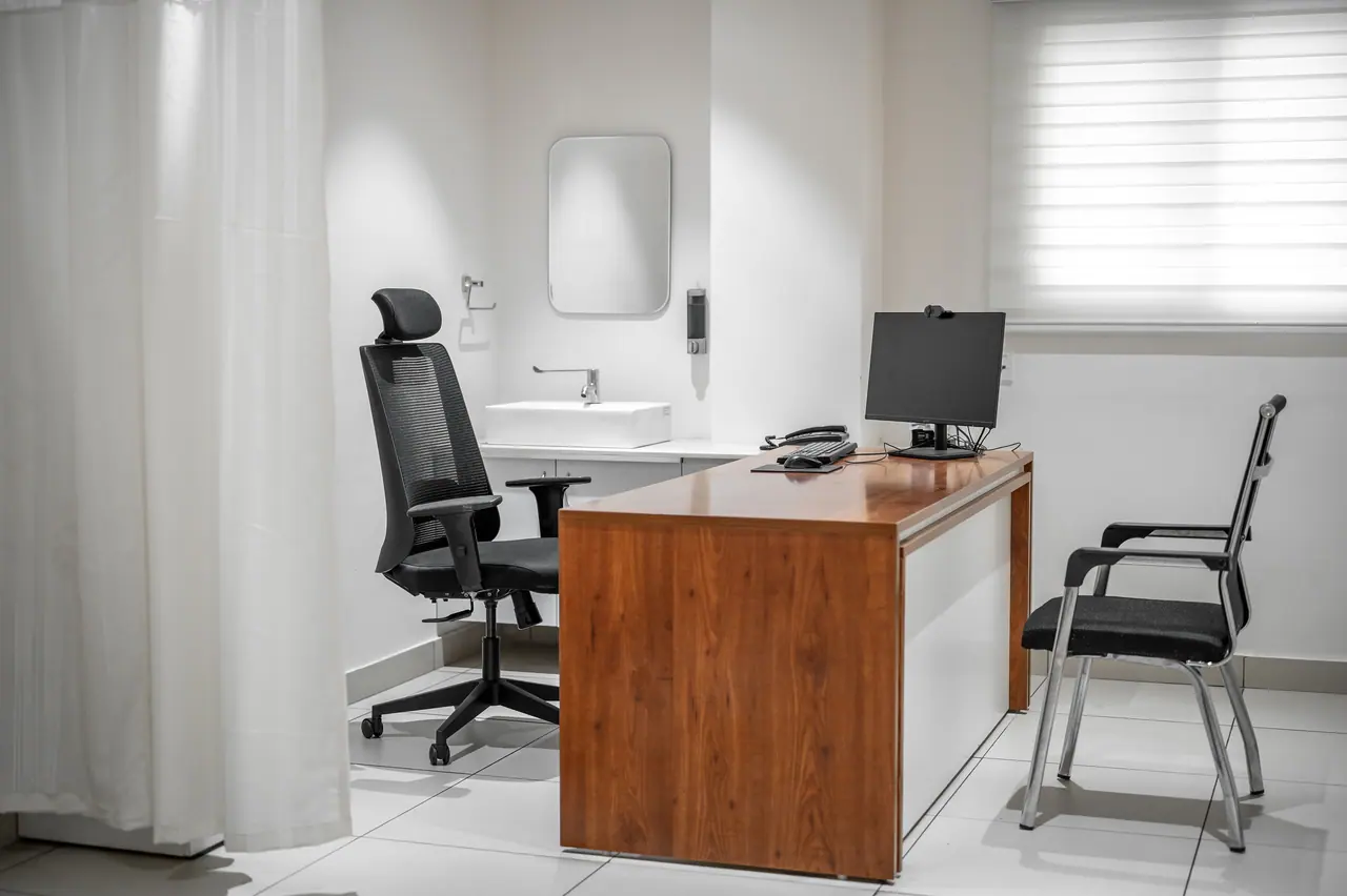 Consultation room with a wood-veneer desk, black mesh office chair, monitor, wall basin with mirror and pale window blind