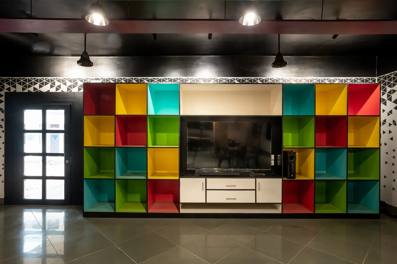 Wall of red, yellow, teal and green open cubbies framing a mounted TV and white drawers, beside a glazed black door