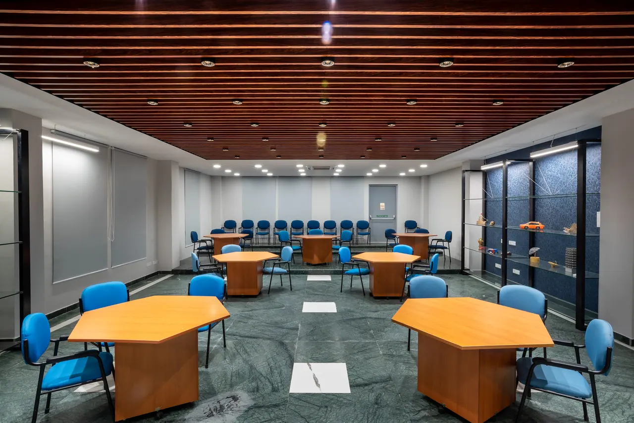 Activity room with hexagonal wooden tables, blue chairs, a wood-slat ceiling, green stone floor and lit glass display shelves
