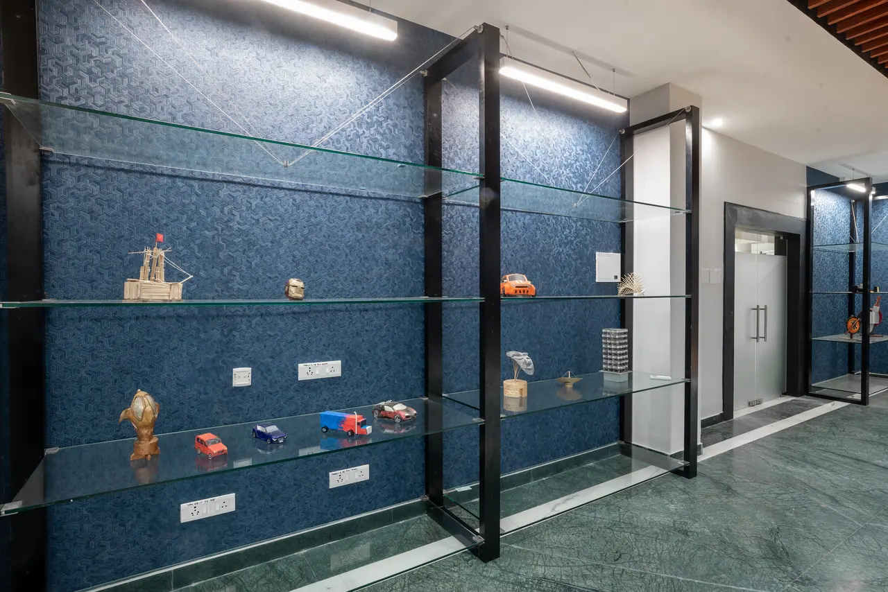 Glass shelves on black steel frames against blue textured wallpaper, holding model cars, a ship model and small sculptures