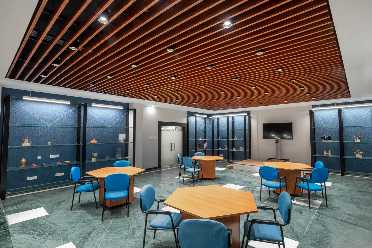 Room under a wood-slat ceiling with blue-chaired hexagonal tables, a raised timber platform, TV on a stand and glass shelves