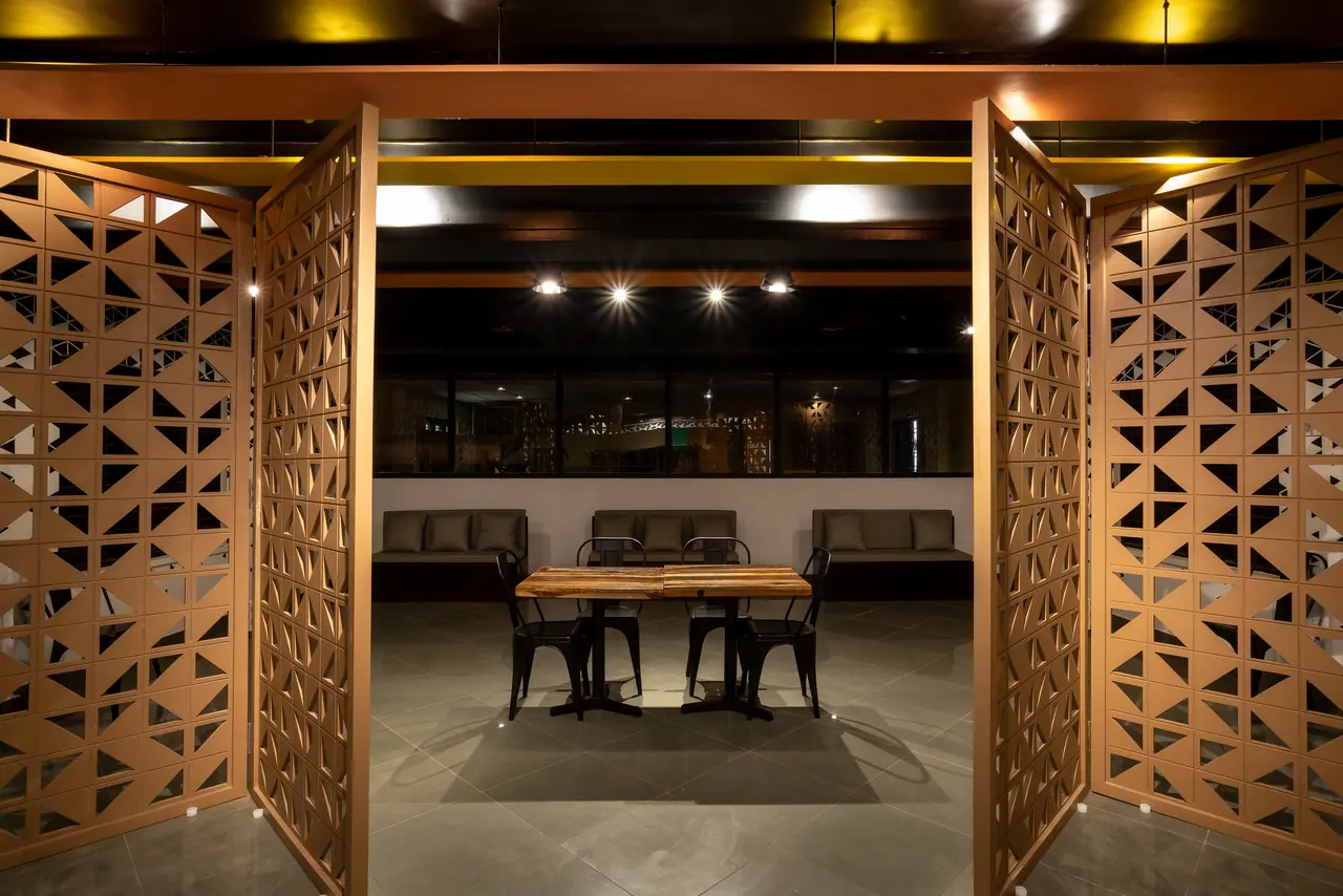 Hinged laser-cut wooden screen panels opening onto a wooden table, black metal chairs and upholstered banquettes beyond