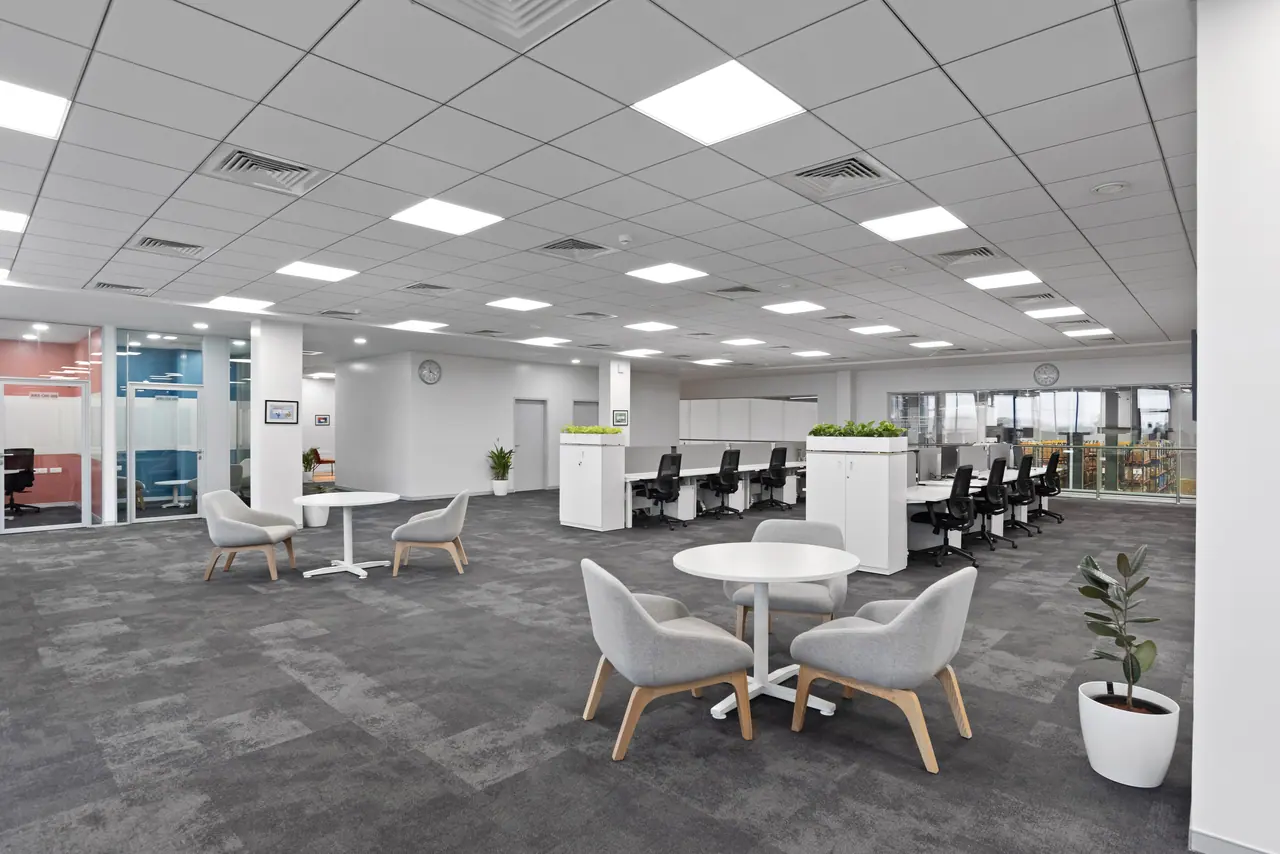 Open-plan office with grey lounge chairs around round white tables, white workstations and glass meeting rooms