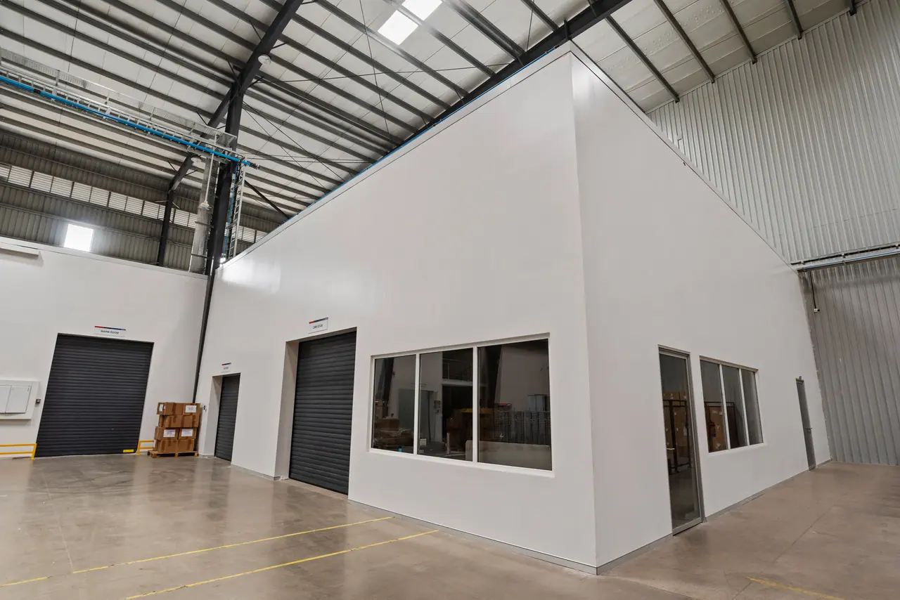 Warehouse hall with a white-panelled room block, black roller shutters and windows under a steel truss roof