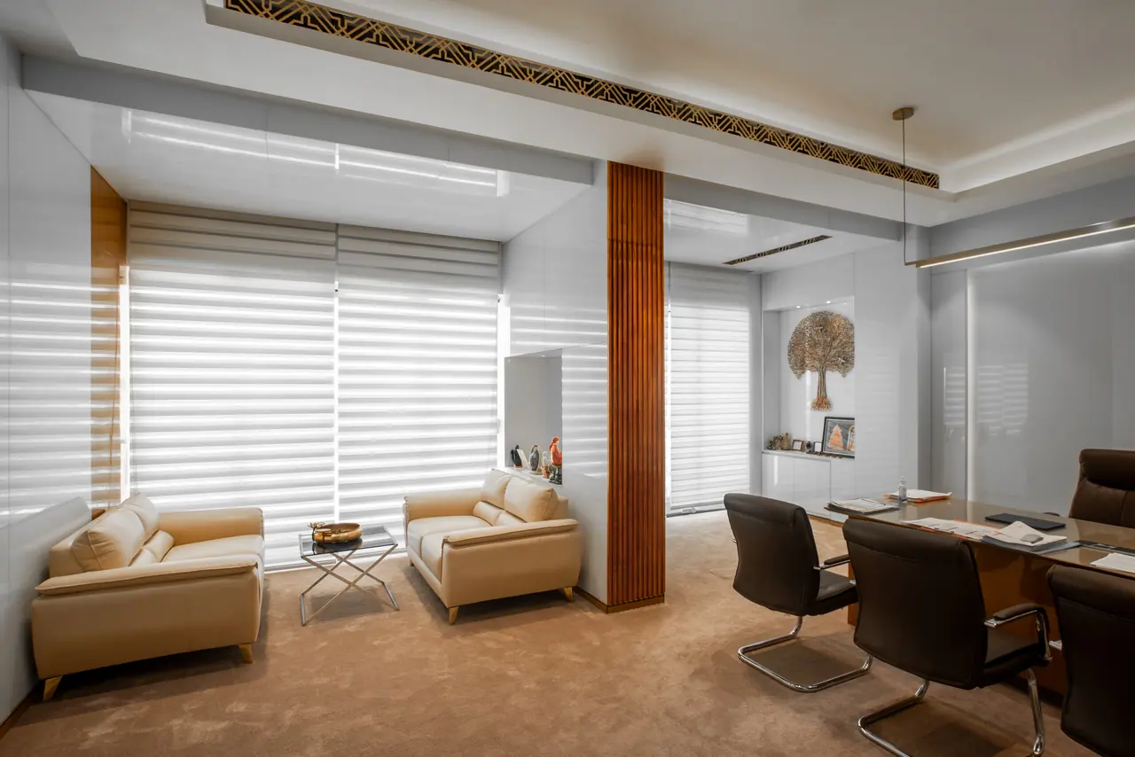 Office seating corner with two cream sofas, a glass side table and a slatted wood column beside light-filtering blinds