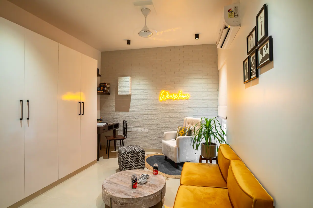 Lounge room with tall white wardrobes, a mustard velvet sofa, a white brick wall with yellow neon script and a study desk