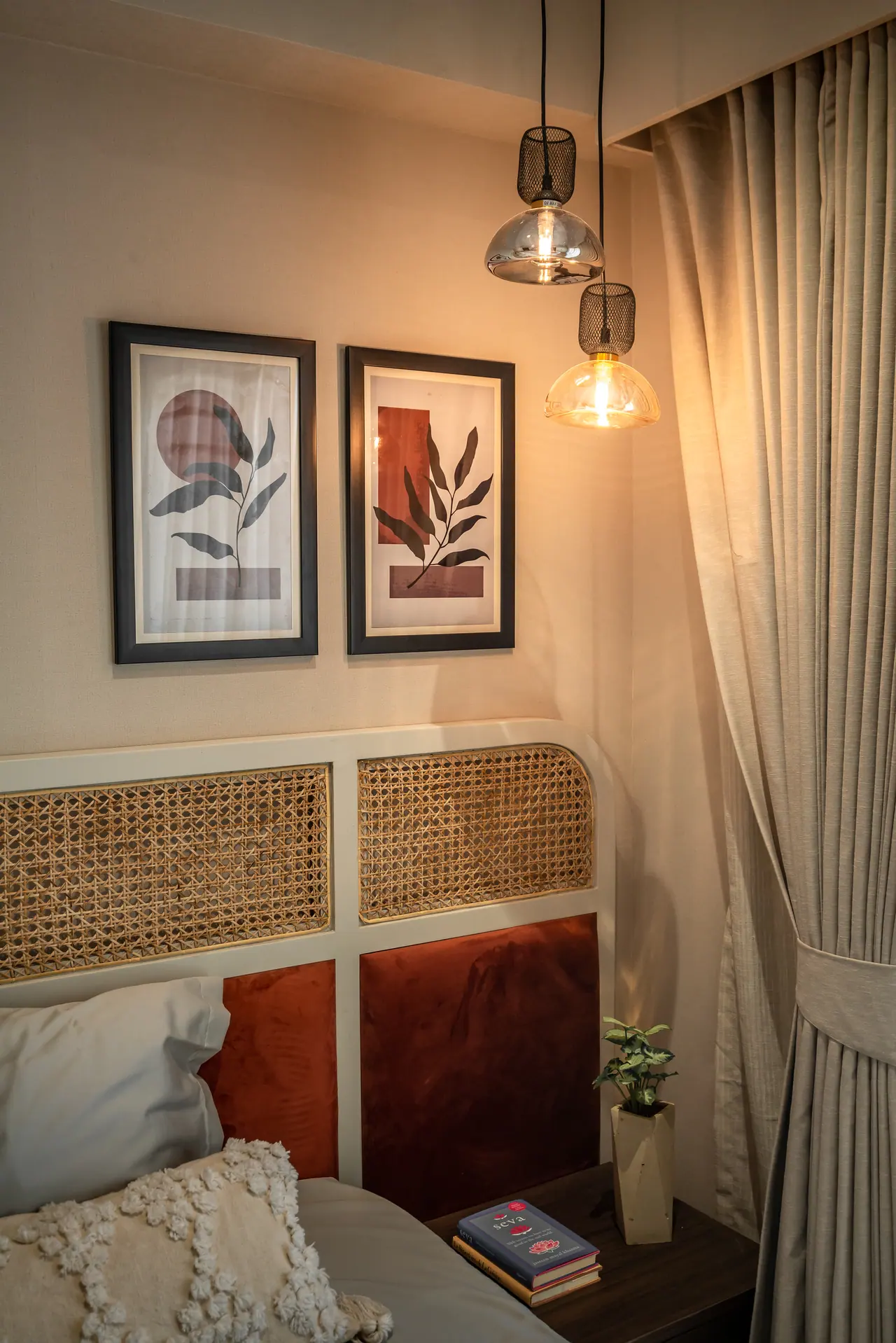 Cane and rust velvet headboard beneath two framed leaf prints, with glass pendant lights beside a beige curtain