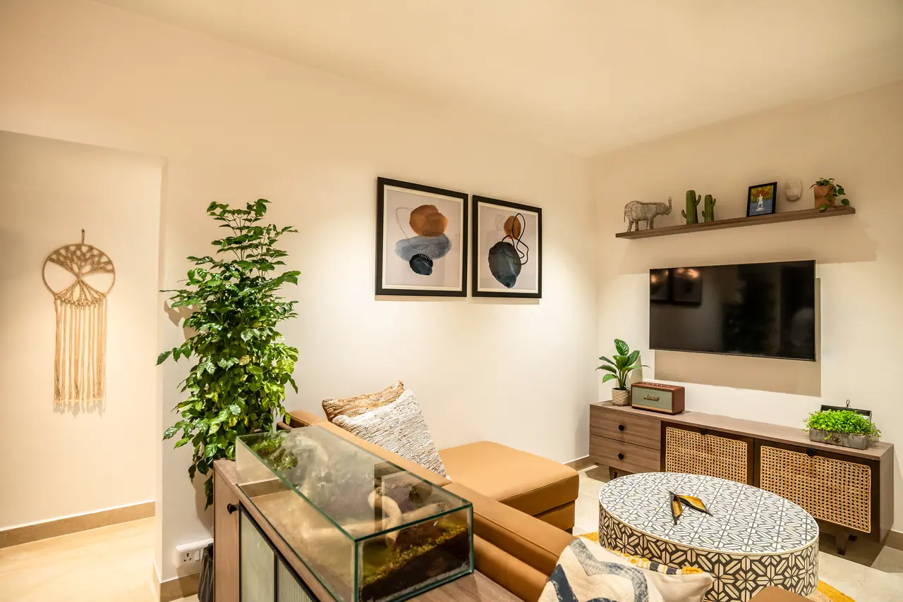 Living area with a tan sofa, patterned drum coffee table, cane-front TV unit, abstract prints and a tall potted plant