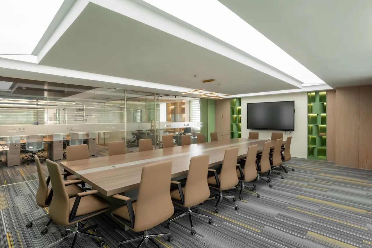 Boardroom with a long walnut table, tan leather chairs, a wall screen, backlit green shelving and glass partitions