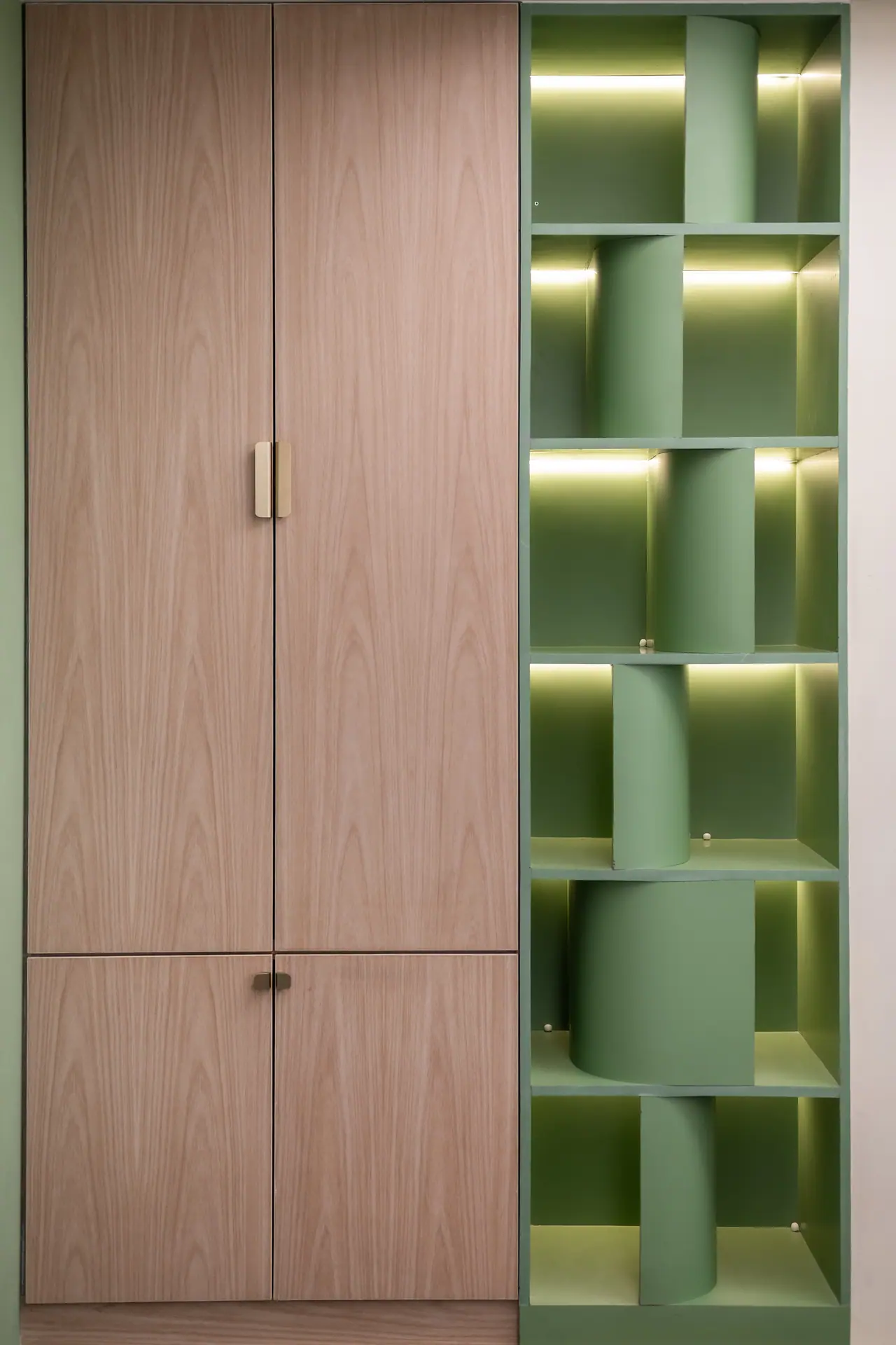 Tall wood-veneer cabinet with brass handles beside lit green open shelving with curved green inserts