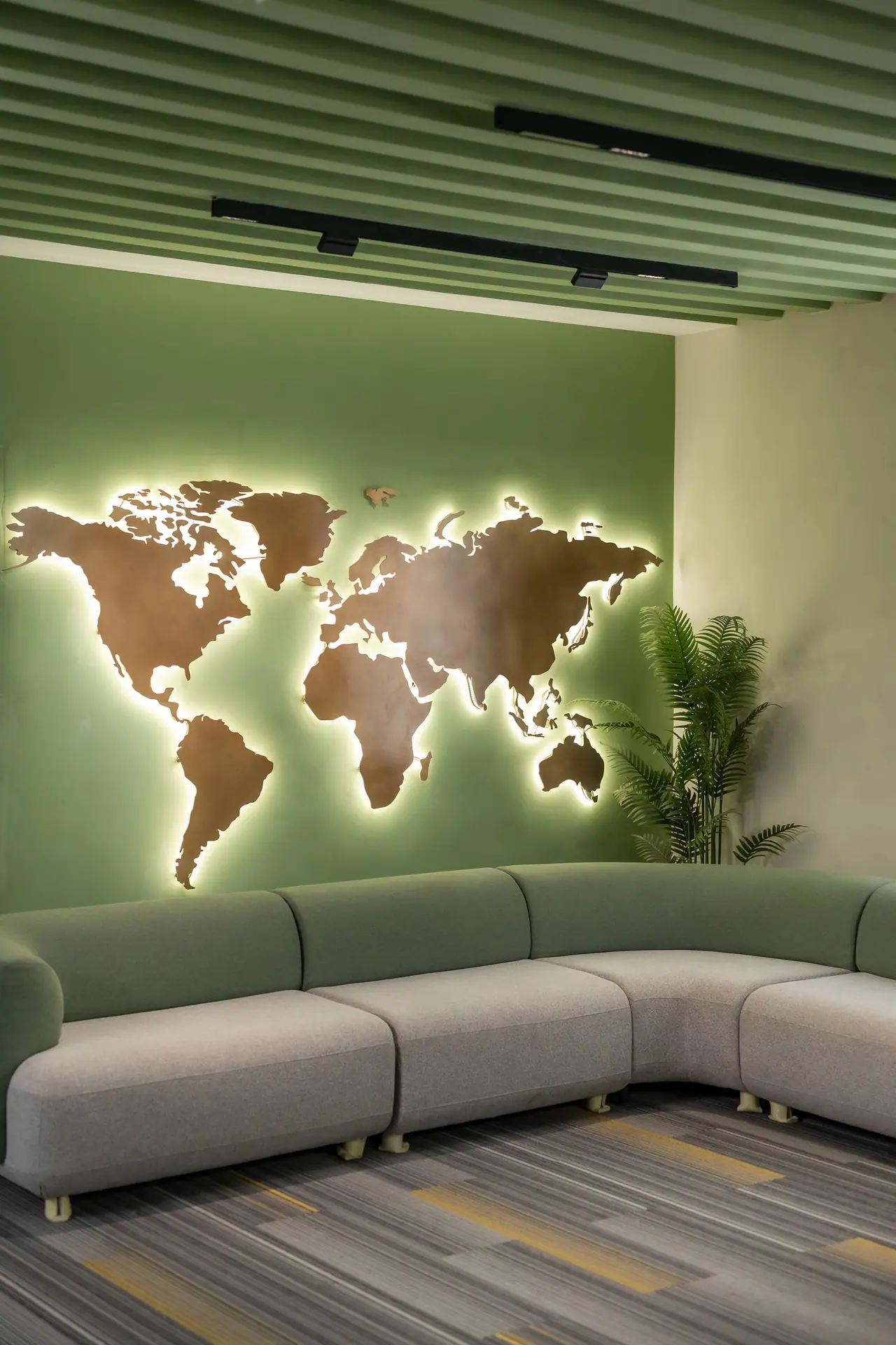 Backlit world map cut-out on a green wall above a curved grey sofa, under a green slatted ceiling and beside a palm