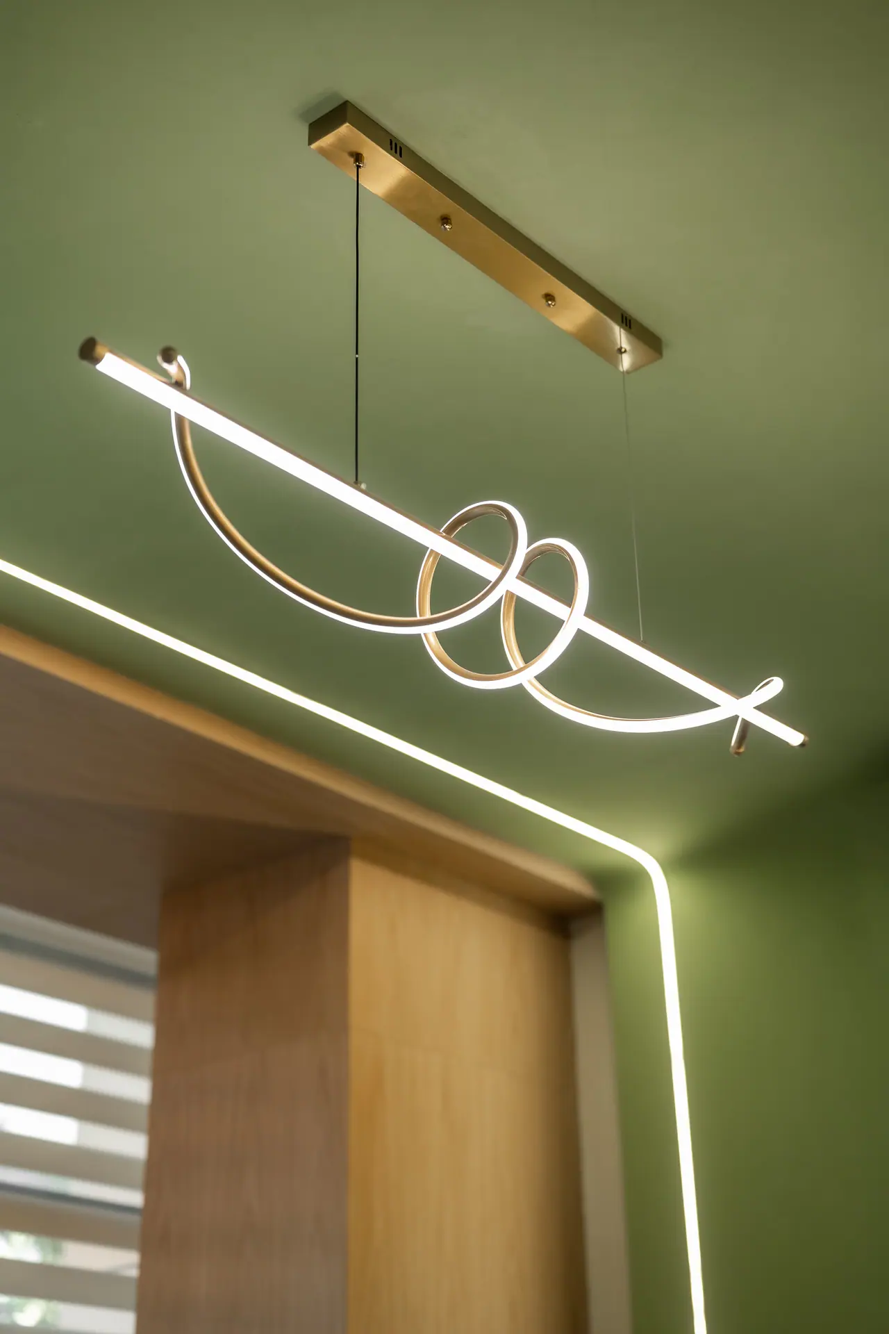 Brass looped linear pendant light on a green ceiling, with a glowing light strip tracing an oak-lined window reveal