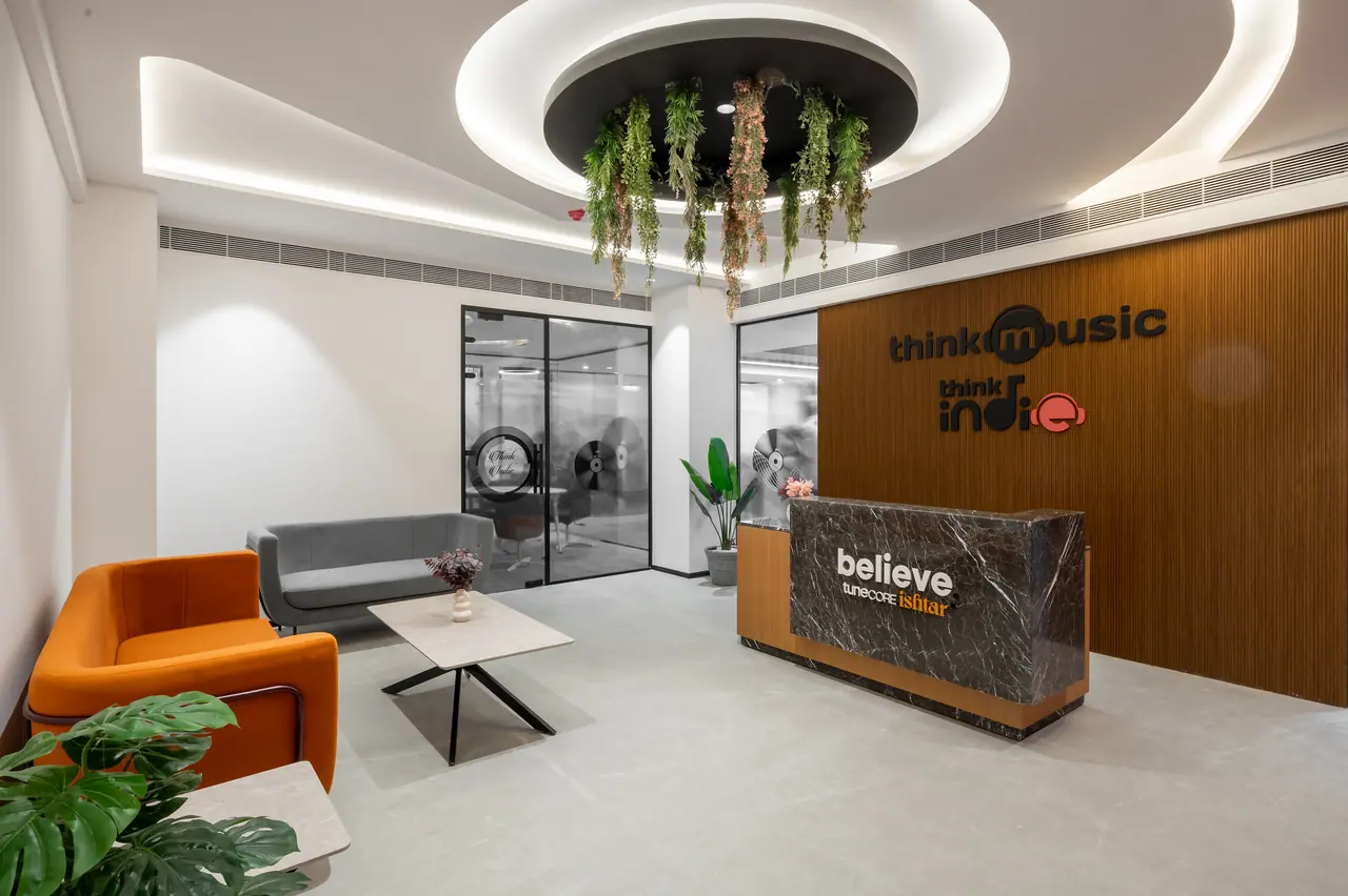 Reception with a dark marble desk, orange armchair, grey sofa, wood-slat logo wall and a lit circular ceiling with greenery
