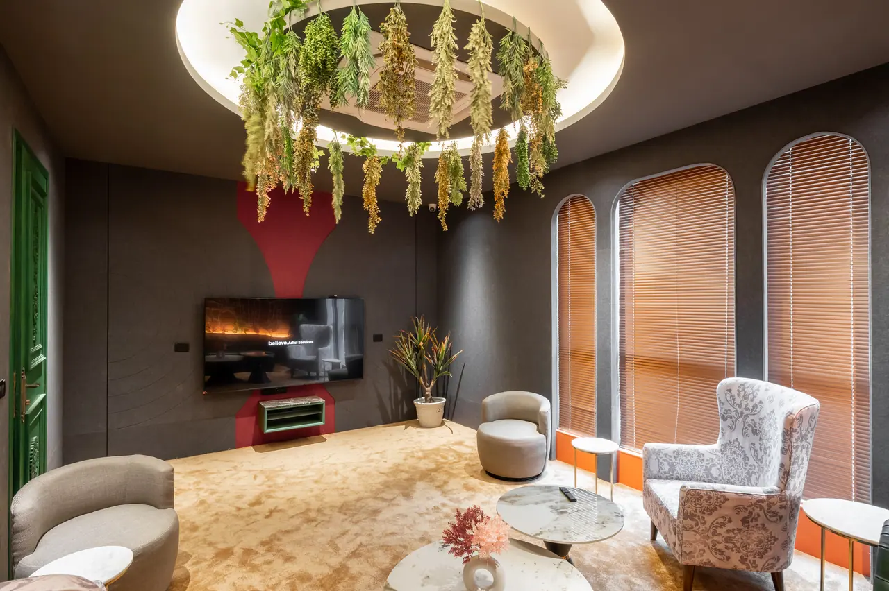 Lounge with tall arched windows and blinds, a wall-mounted TV on a red accent, hanging dried foliage and a gold-toned carpet
