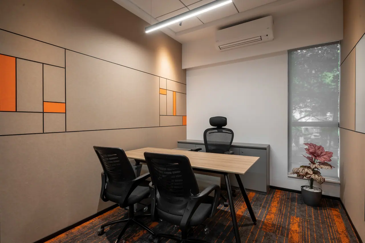 Small cabin with a light wood desk, black mesh chairs, beige wall panels with orange accents and a tall window with a blind