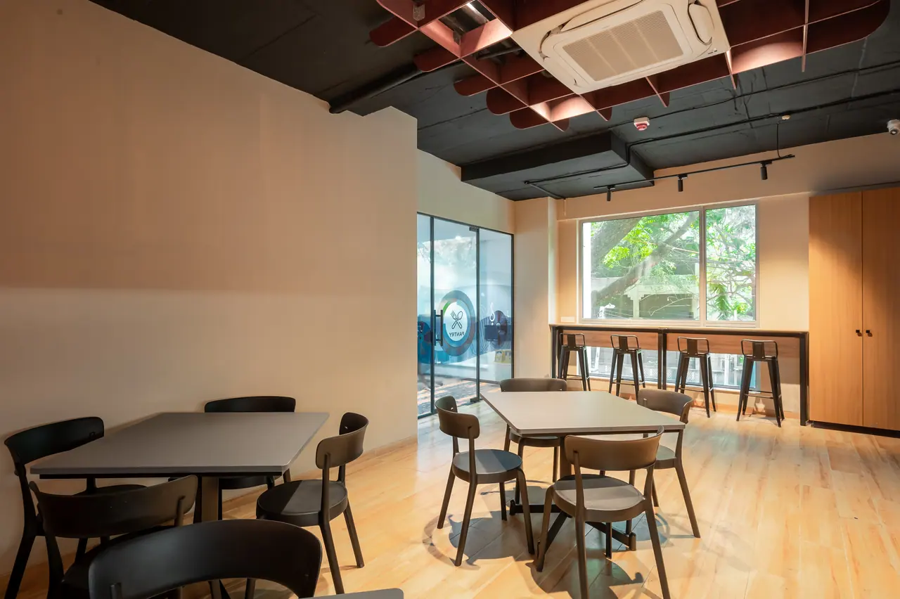 Cafeteria with dark tables and chairs on wood-look flooring, a red baffle ceiling and a window counter with metal bar stools