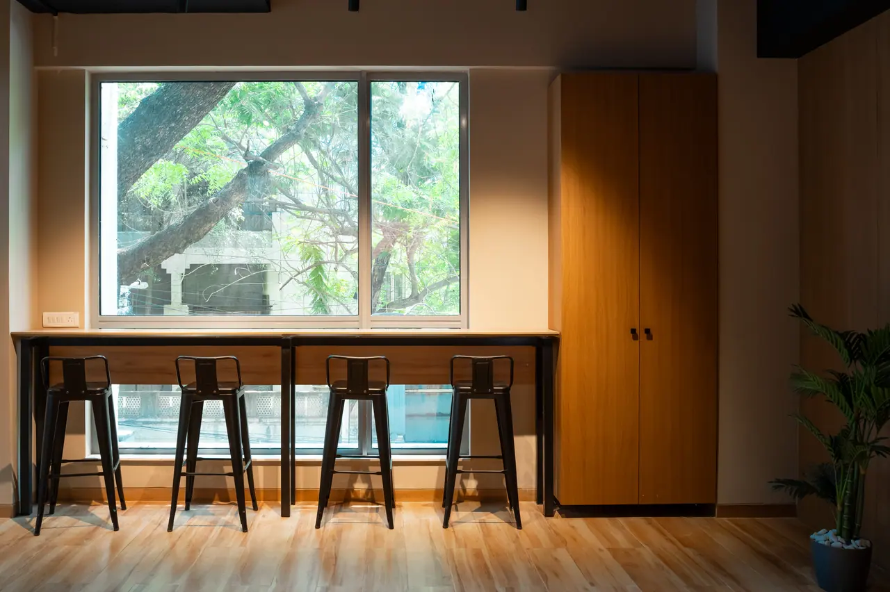 Window counter with four black metal bar stools looking out to tree branches, beside a tall wood-finish cabinet