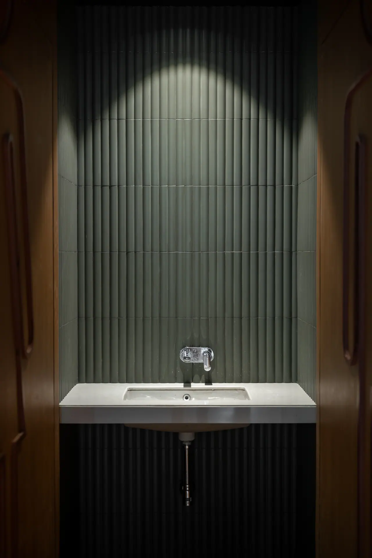 Washbasin niche lined with fluted green tiles under a single spotlight, framed by curved wood-panelled doors