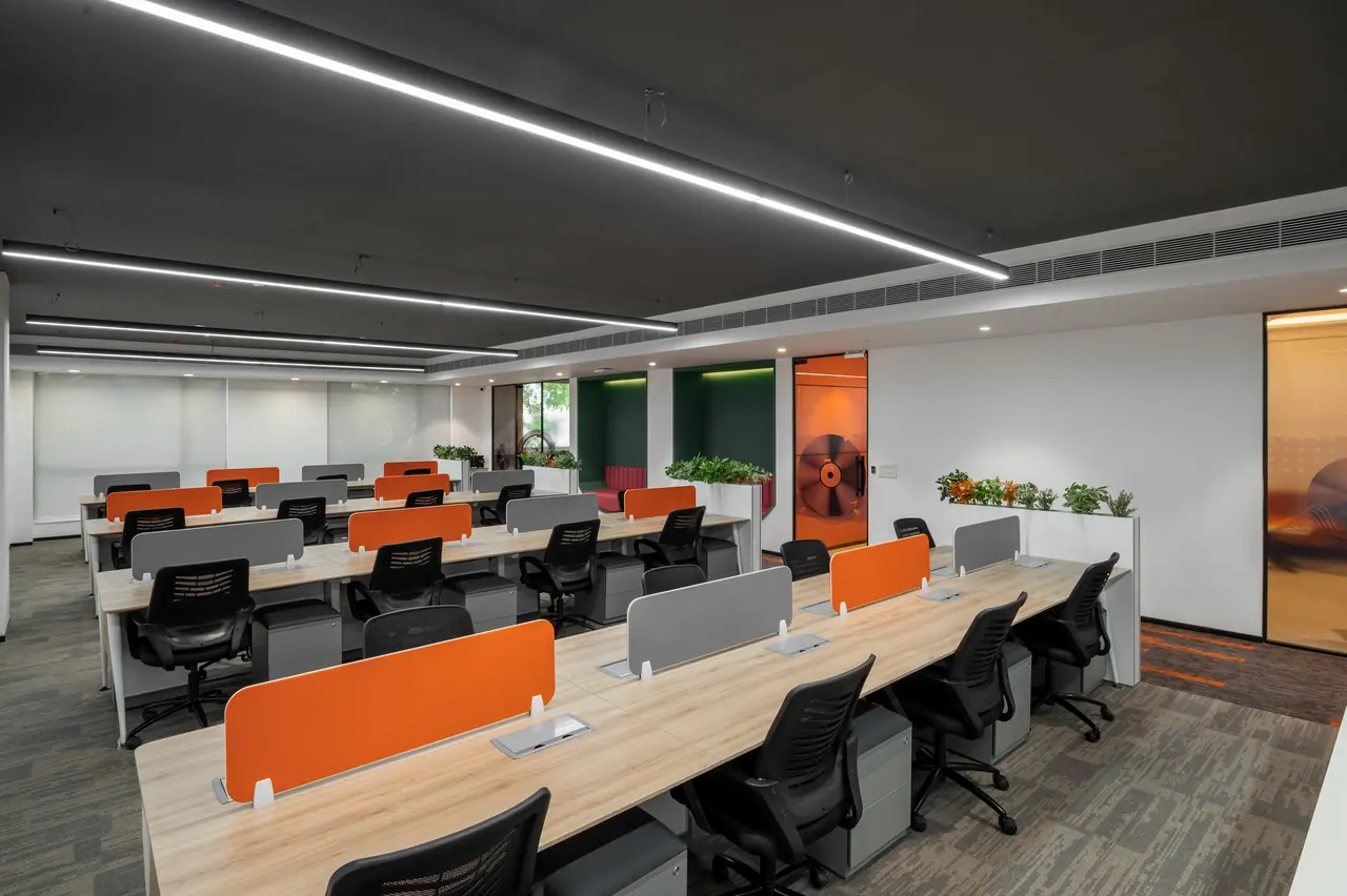 Open-plan workstations with orange and grey desk screens under linear lights and a dark ceiling, green niche seats beyond