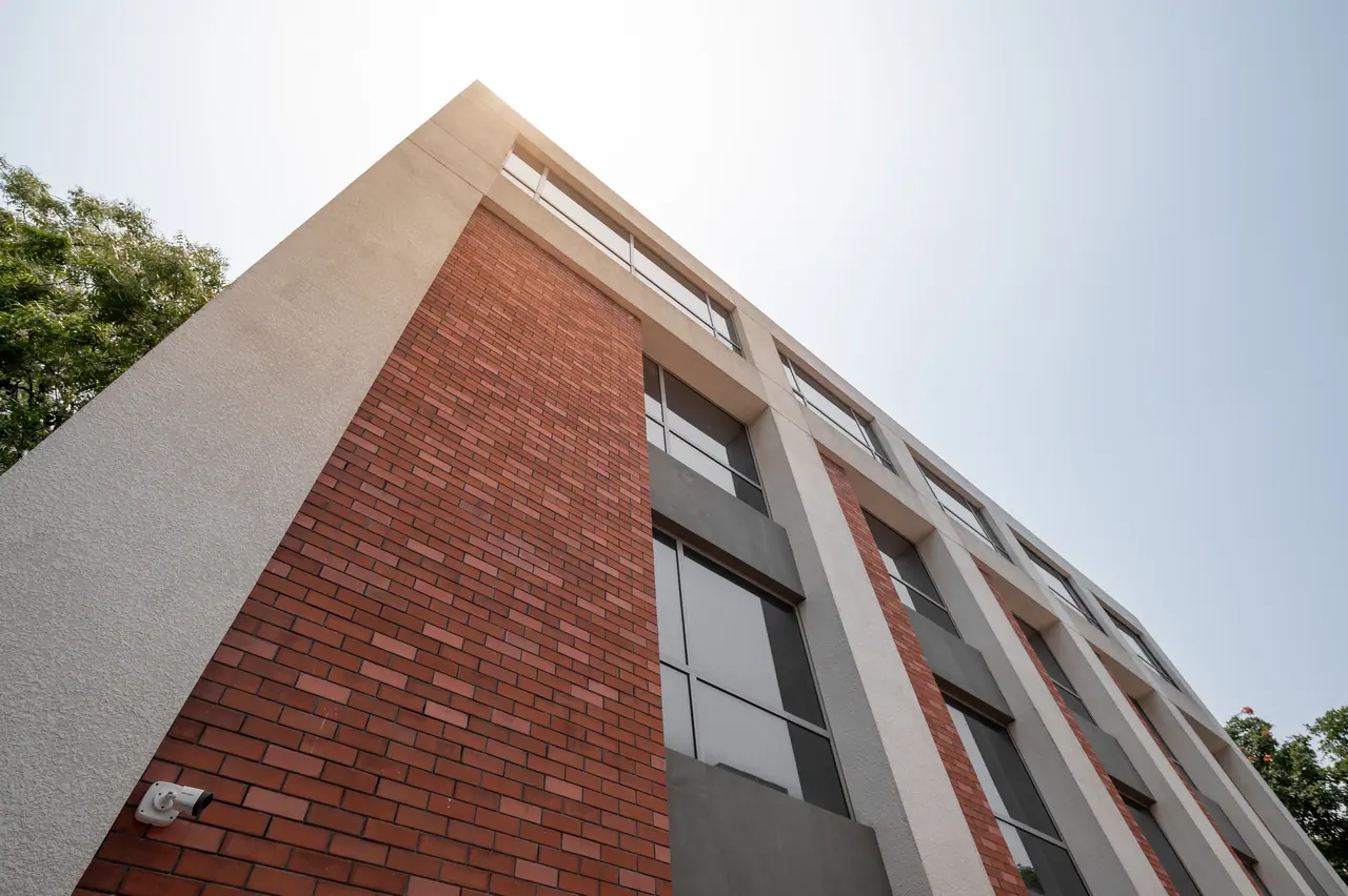 Low-angle view of a multi-storey facade with red brick panels, textured render frames and tall windows against a pale sky
