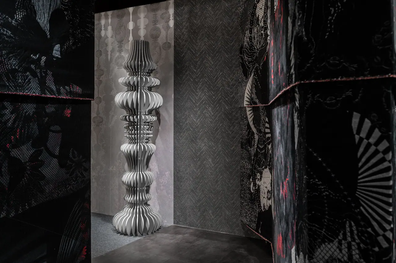 Dark room with a tall pleated white column sculpture framed by black wall panels printed with red-accented line art