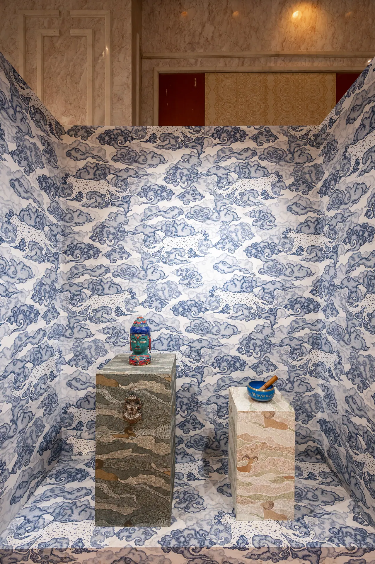 Booth wrapped in blue cloud-pattern wallcovering, with patterned plinths holding a painted head sculpture and a bowl