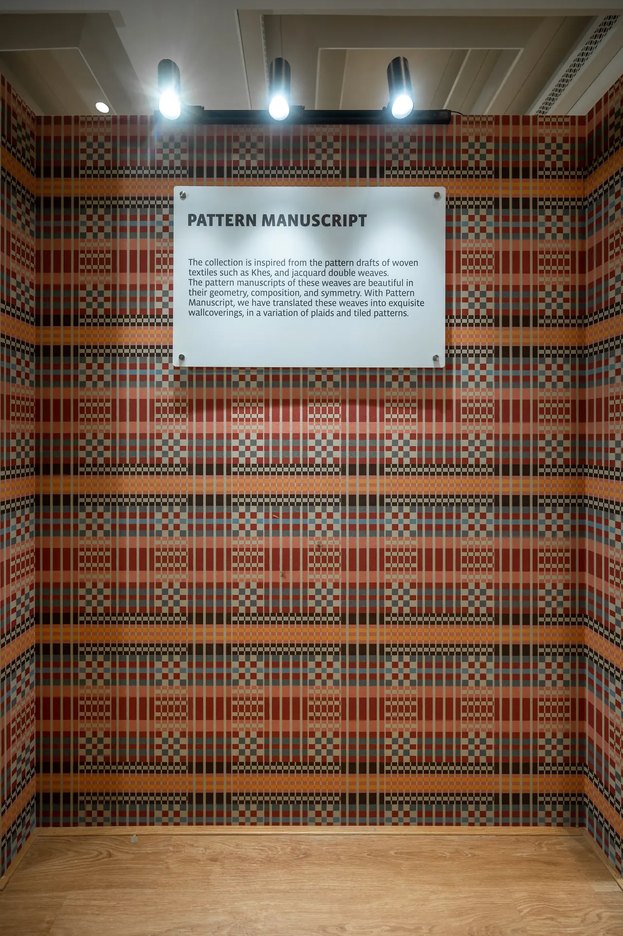 Booth lined in red, orange and grey woven-plaid wallcovering under three spotlights, with a white text panel on the wall