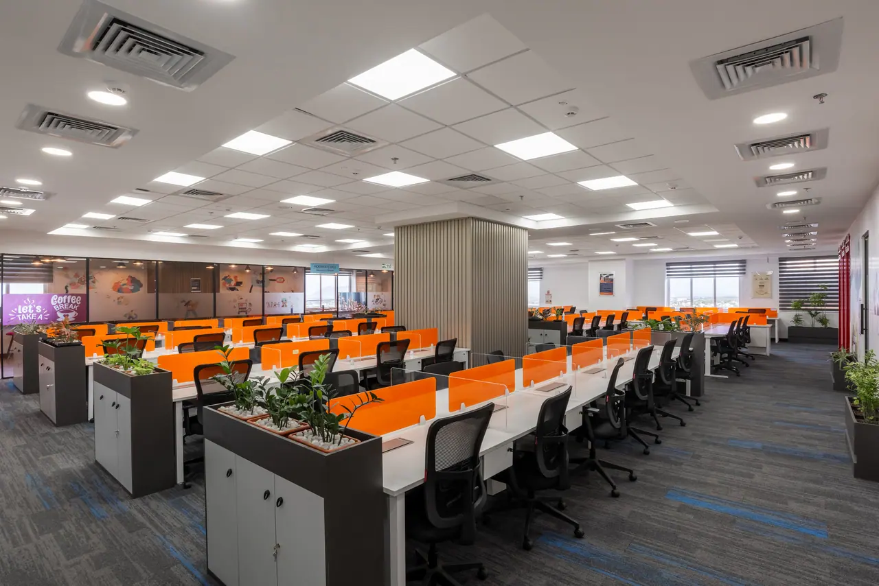 Rows of workstations with orange desk screens and grey planter cabinets around a slatted column under a white grid ceiling