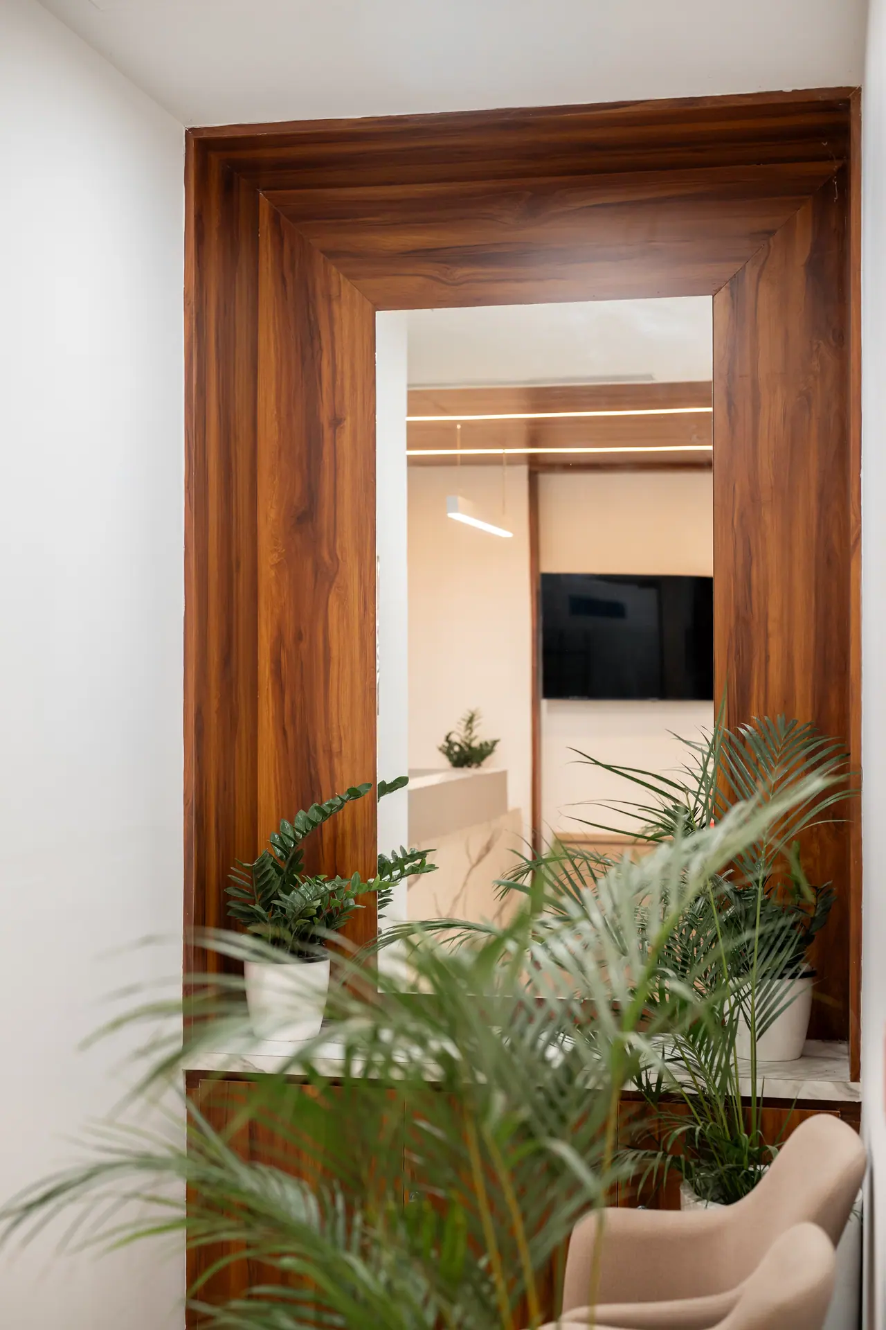 Tall wood-veneer framed mirror reflecting the room beyond, with potted palms and a beige armchair in the foreground
