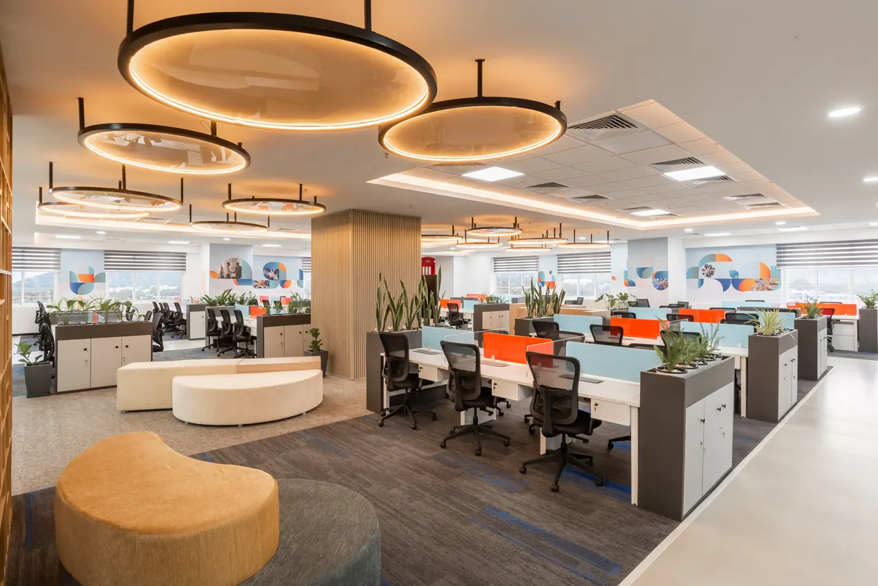 Open office with curved upholstered seating beneath large circular pendant rings, desk clusters and a slatted wood column