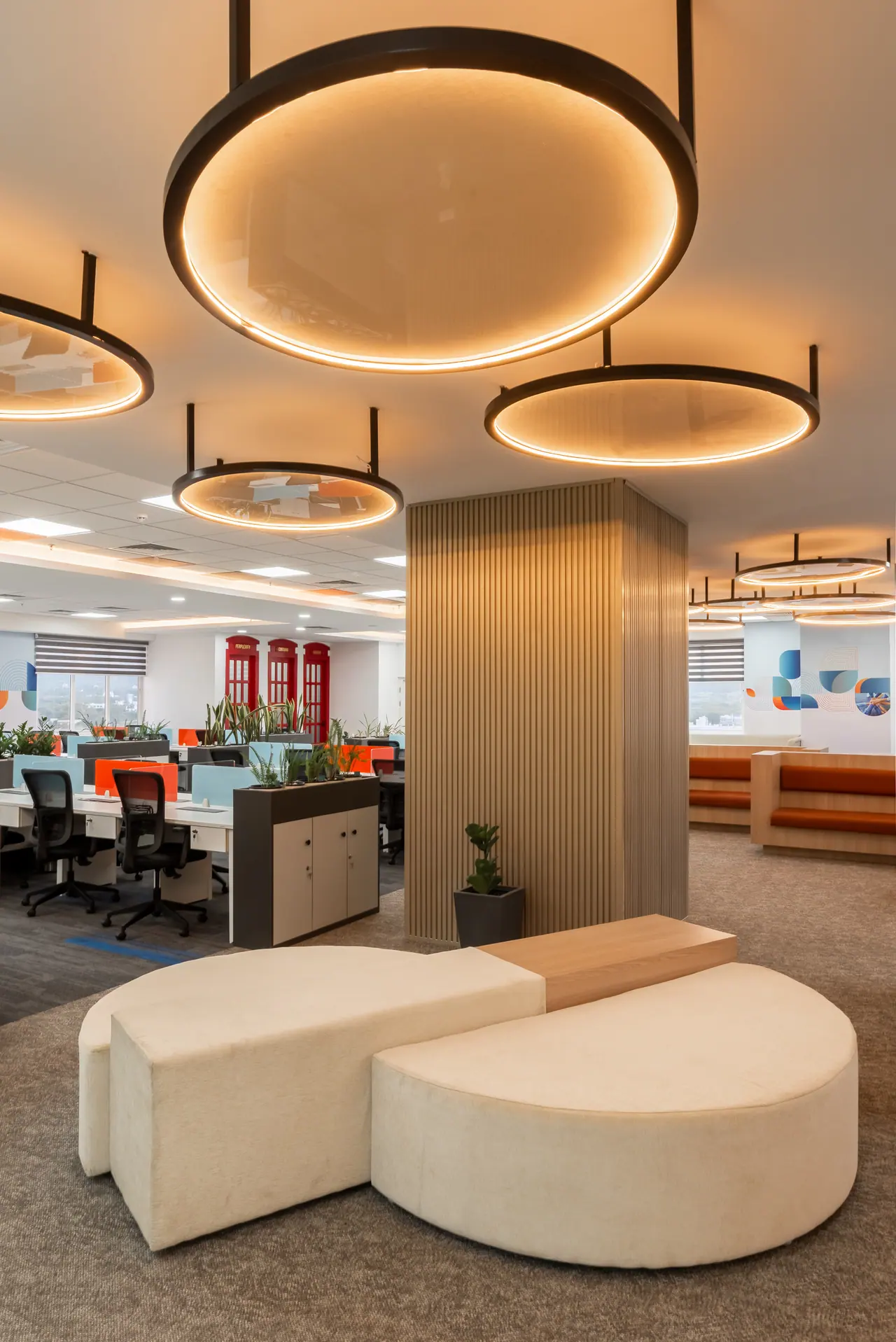 Cream upholstered curved bench seating below glowing circular pendant rings beside a slatted column in an open office